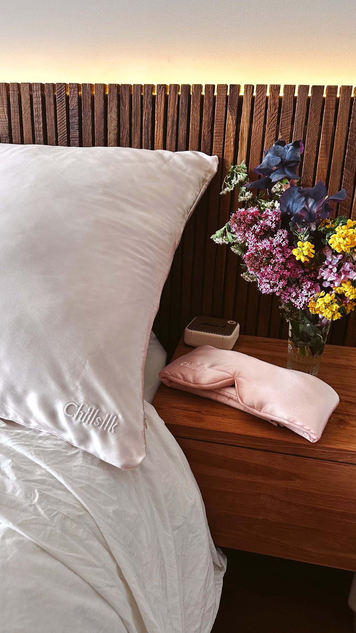 Taie d'oreiller en soie couleur rose blush, un rose très pâle. Une tête de lit en bois avec un néon lumineux derrière. une table de chevet avec un bouquet de fleurs, une enceinte et un masque de nuit en soie couleur rose clair.  Image